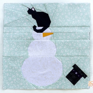 May include: A snowman with a black cat sitting on top of its head. The snowman is made of white fabric and has a yellow carrot nose. The background is a light blue fabric with white polka dots.