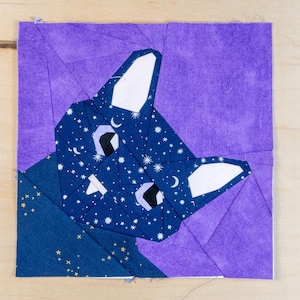 Może przedstawiać: Praca z tkaniny przedstawiająca stylizowaną twarz kota. Twarz kota to patchwork z niebieskiej tkaniny z wzorem gwiaździstej nocy, białymi uszami i czarnymi oczami. Tło jest jednolite fioletowe.