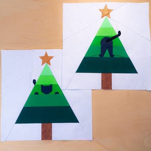 May include: Two fabric blocks featuring Christmas trees. Each tree is green with a brown trunk, a gold star, and a dark blue cat. One cat is peeking out, the other is showing its rear. The trees are on a white background.