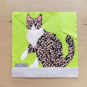 以下が含まれることがあります： 黒い花柄の猫が描かれた、ライムグリーンの背景のパッチワークブロック。猫は白いアクセントとピンクの耳を持っています。正方形のブロックは明るい木製の表面に置かれています。