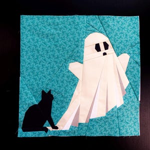 May include: A square fabric quilt block featuring a teal background with a small floral pattern. A white ghost with black eyes and a black mouth is positioned on the right, while a solid black cat sits on the left, facing toward the ghost.