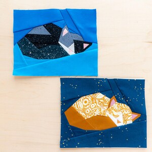 May include: Two fabric art pieces with a sleeping fox design. One fox is grey and black on a starry background, the other is orange and yellow on a dark blue background. The pieces are made of geometric shapes and are square in form.