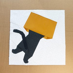 Può includere: Un'opera d'arte geometrica in tessuto raffigurante un gatto nero con una scatola gialla sulla testa. Il gatto è fatto di tessuto nero fantasia e la scatola è giallo pieno. Lo sfondo è bianco con piccoli punti grigi.