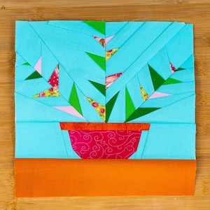 Pode incluir: Uma aplicação de tecido azul de uma planta em um vaso rosa com um caule verde e flores rosa e amarelas. A planta está sobre um fundo laranja claro.
