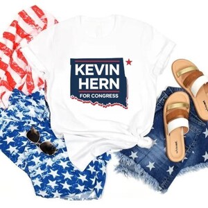 Puede incluir: Camiseta blanca con un gráfico azul y rojo que dice "Kevin Hern for Congress" con una estrella roja y un contorno de Oklahoma. La camiseta se combina con pantalones cortos de mezclilla con estrellas blancas y sandalias marrones.
