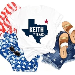 May include: White t-shirt with a blue outline of the state of Texas and the text "Keith for Texas" in red and blue. A red star is above the state outline. The t-shirt is laid out on a white background with blue and red star-patterned fabric and denim shorts.
