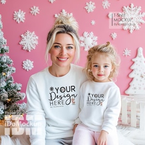 Op de afbeelding: Twee mensen dragen witte sweatshirts met de tekst "YOUR DESIGN HERE" en een rendier afbeelding. De sweatshirts zijn op een roze achtergrond met witte sneeuwvlokken.