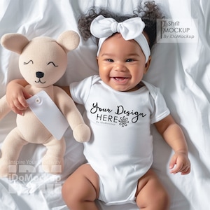 May include: A white baby onesie with the text "Your Design HERE" and a small flower graphic. The onesie is being worn by a baby with dark curly hair and a white headband with a bow. The baby is lying on a white sheet and holding a beige teddy bear.