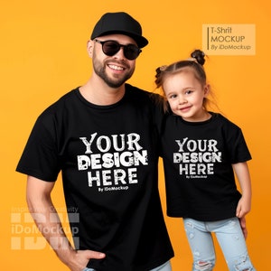 May include: Black t-shirts with the text "YOUR DESIGN HERE" in white, worn by an adult and a child. The adult wears a black cap and sunglasses. The child wears blue jeans. The background is orange.