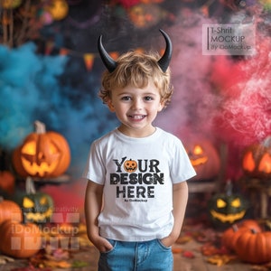 May include: A young child wearing a white t-shirt with the text "YOUR DESIGN HERE" and a pair of black devil horns. The child is standing in front of a Halloween-themed background with pumpkins and smoke.