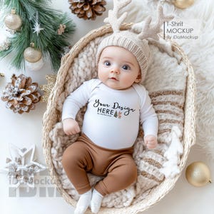 Op de afbeelding: Een witte baby romper met lange mouwen met de tekst "Your Design Here" en een kleine afbeelding van bomen. De romper wordt gedragen door een baby die in een rieten mand ligt met een bruin-witte deken. De baby draagt een beige gebreide muts met rendiergewei.