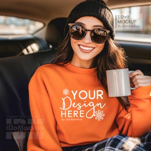 Könnte beinhalten: Eine Frau mit schwarzer Mütze und Sonnenbrille sitzt in einem Auto und hält eine weiße Tasse. Sie trägt ein orangefarbenes Sweatshirt mit dem Text "YOUR Design HERE" und einem weißen Blumenmotiv.