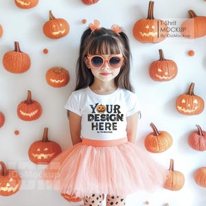 May include: A young girl wearing a white t-shirt with the text "YOUR DESIGN HERE" printed on it. She is wearing a pink tulle skirt and sunglasses. The background is a white wall with orange pumpkins with jack-o-lantern faces.