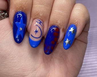 May include: A hand displaying a manicure with blue and nude-colored nail polish. The nails feature star and abstract designs in silver and blue. One nail has a blue star with a silver gem in the center.