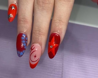 May include: Close-up of a hand with long, oval-shaped nails. The nails are painted in various designs, including red with star accents, a blue and red pattern, a pink nail with a crescent moon and star design, and a red star with a rhinestone.