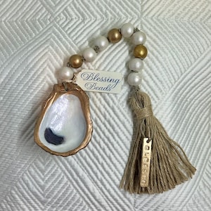 May include: A gold and white oyster shell charm with a tassel and beads. The charm has the word "Blessed" on it and the beads have a tag that says "Blessing Beads".