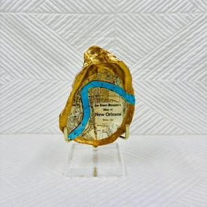 May include: A gold-painted oyster shell with a map of New Orleans, featuring a turquoise river. The shell is mounted on a clear acrylic stand. The text on the map reads "The Times Picayune's Map of New Orleans."