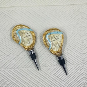 May include: Two bottle stoppers with a gold-colored, ornate design. Each stopper features a map of New Orleans with a blue ribbon accent. The stoppers have a black rubber seal and a silver pointed tip. The background is a white, textured surface.