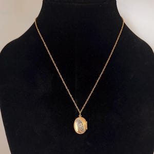 May include: A gold-colored oval locket necklace displayed on a black mannequin. The locket has a small, decorative design on the front. The necklace chain is delicate and gold-toned.
