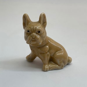 May include: A small, beige ceramic figurine of a French bulldog sitting with its ears perked up.
