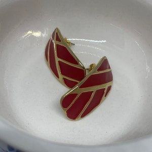 May include: A pair of red and gold-toned stud earrings. The earrings have a leaf-like shape with a red enamel finish and gold-colored outlines. The posts are gold-colored.