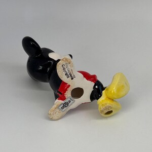 Puede incluir: Una figura de cer&aacute;mica de Mickey Mouse, vista desde abajo. La figura es principalmente negra, con pantalones cortos rojos y zapatos amarillos. La base es blanquecina, con el texto "Disney Productions Copyright".