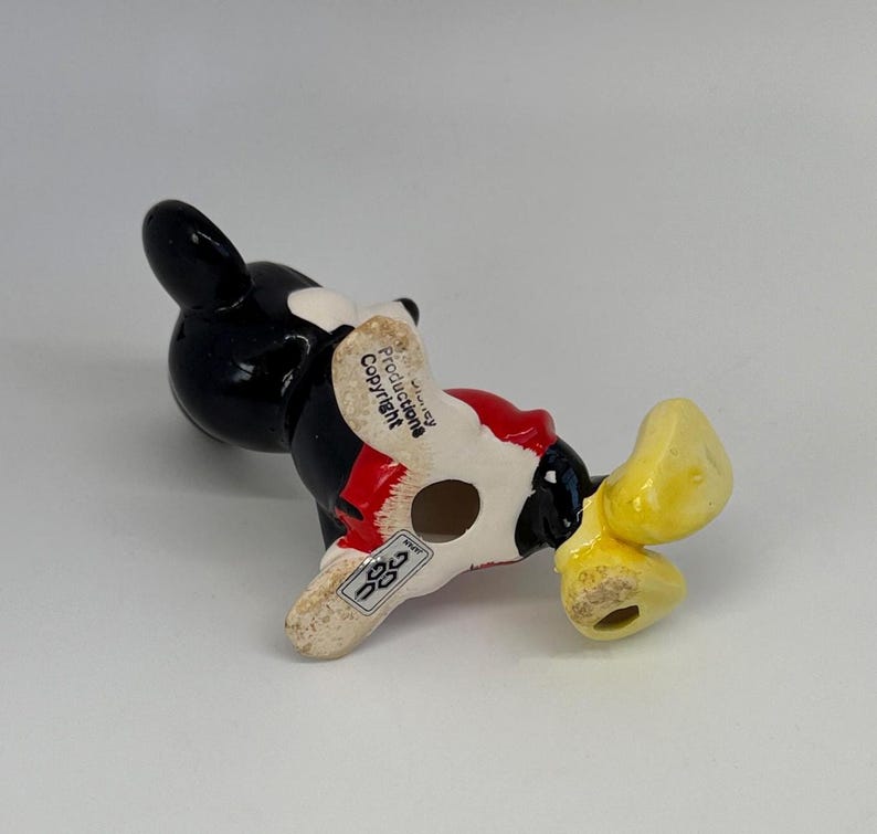Puede incluir: Una figura de cer&aacute;mica de Mickey Mouse, vista desde abajo. La figura es principalmente negra, con pantalones cortos rojos y zapatos amarillos. La base es blanquecina, con el texto "Disney Productions Copyright".