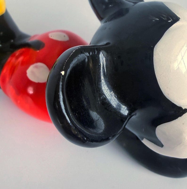 Puede incluir: Primer plano de una figura vintage de Mickey Mouse. La figura presenta una cabeza y una oreja negras, una cara blanca y un cuerpo rojo con lunares blancos. La figura es brillante y parece estar hecha de cer&aacute;mica o un material similar.