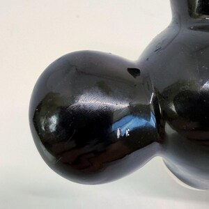 Op de afbeelding: Close-up van een glanzend zwart keramiek beeldje. Het beeldje heeft een afgeronde vorm met gladde, reflecterende oppervlakken. De details zijn minimaal, met de nadruk op de vorm en het spel van licht op het zwarte glazuur. Het beeldje lijkt een decoratief item te zijn.