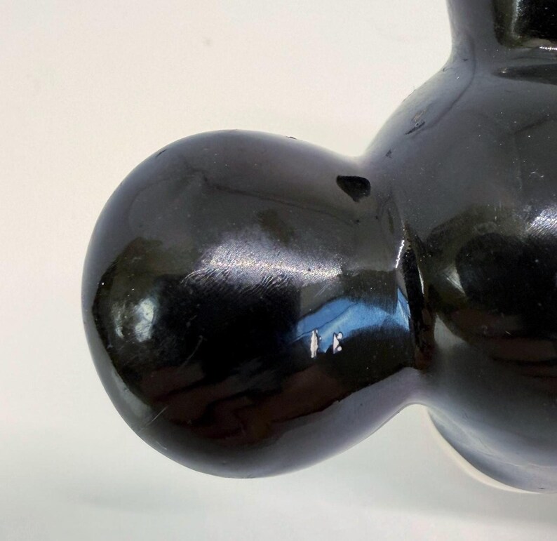 Op de afbeelding: Close-up van een glanzend zwart keramiek beeldje. Het beeldje heeft een afgeronde vorm met gladde, reflecterende oppervlakken. De details zijn minimaal, met de nadruk op de vorm en het spel van licht op het zwarte glazuur. Het beeldje lijkt een decoratief item te zijn.