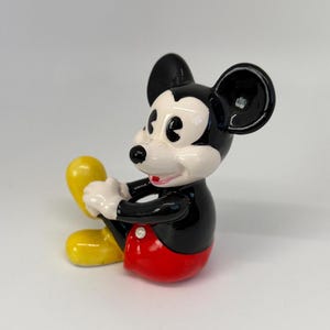 Op de afbeelding: Een keramieken beeldje van Mickey Mouse in een zittende positie. Het figuur heeft een zwart lichaam, een wit gezicht en een rode broek. De oren zijn zwart en de schoenen geel. Een klassiek Disney-personage.