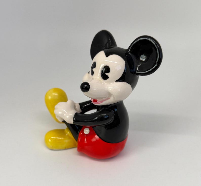 Op de afbeelding: Een keramieken beeldje van Mickey Mouse in een zittende positie. Het figuur heeft een zwart lichaam, een wit gezicht en een rode broek. De oren zijn zwart en de schoenen geel. Een klassiek Disney-personage.