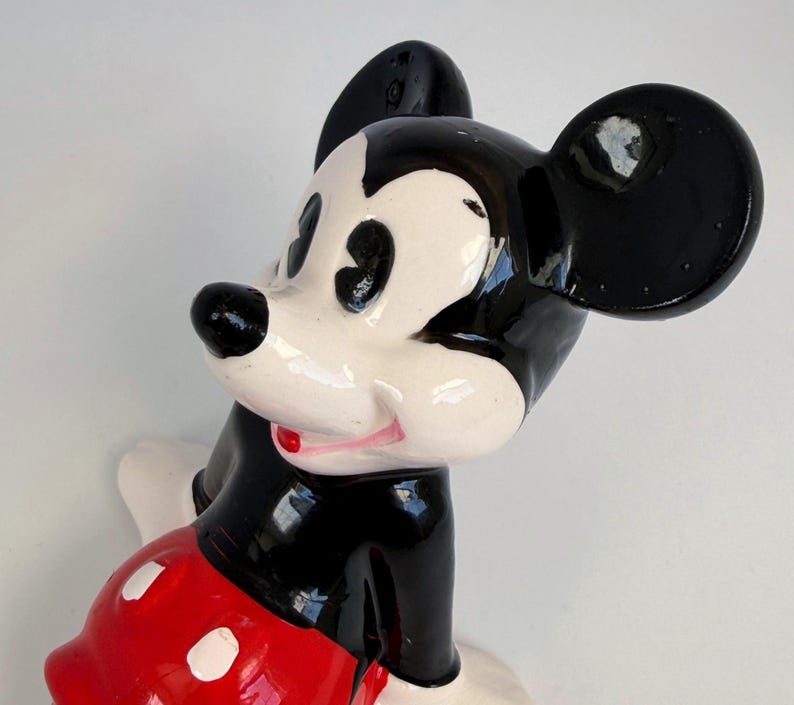 Puede incluir: Una figura de cer&aacute;mica de Mickey Mouse. Tiene un cuerpo, orejas y nariz negros, con una cara blanca y pantalones cortos rojos con botones blancos. La figura tiene un acabado brillante y un dise&ntilde;o de dibujos animados cl&aacute;sico.