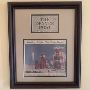 May include: Framed vintage newspaper clippings. The top clipping reads "The Denver Post" and "Home Edition." The bottom clipping is titled "Dawning of United States Space History" and depicts a rocket launch. The frame is dark brown.