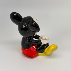 Op de afbeelding: Een keramieken beeldje van Mickey Mouse, met een glanzend zwarte kop en oren, een rode broek, gele schoenen en witte handschoenen. Het beeldje zit met de handen op de knie&euml;n.