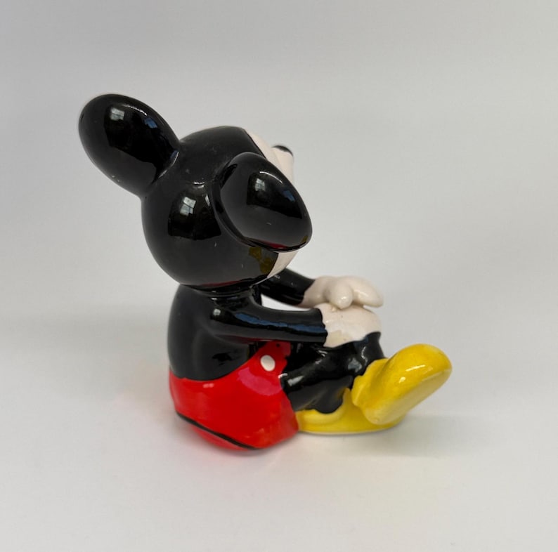 Op de afbeelding: Een keramieken beeldje van Mickey Mouse, met een glanzend zwarte kop en oren, een rode broek, gele schoenen en witte handschoenen. Het beeldje zit met de handen op de knie&euml;n.