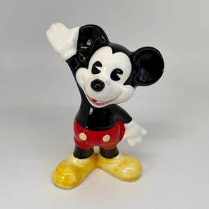 以下が含まれることがあります： 陽気なポーズのミッキーマウスの陶器製フィギュア。フィギュアは黒い体、白い手袋、黄色の靴をしています。ミッキーは白いボタンが付いた赤いショートパンツを履いています。クラシックなディズニーのコレクターアイテムです。