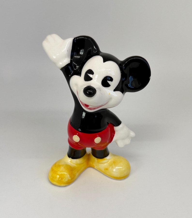 以下が含まれることがあります： 陽気なポーズのミッキーマウスの陶器製フィギュア。フィギュアは黒い体、白い手袋、黄色の靴をしています。ミッキーは白いボタンが付いた赤いショートパンツを履いています。クラシックなディズニーのコレクターアイテムです。