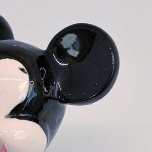 Puede incluir: Primer plano de una figura de Mickey Mouse en blanco y negro. La figura tiene un acabado brillante, con una gran oreja negra y una parte de la cara visible. La cara es blanca con acentos rosas.