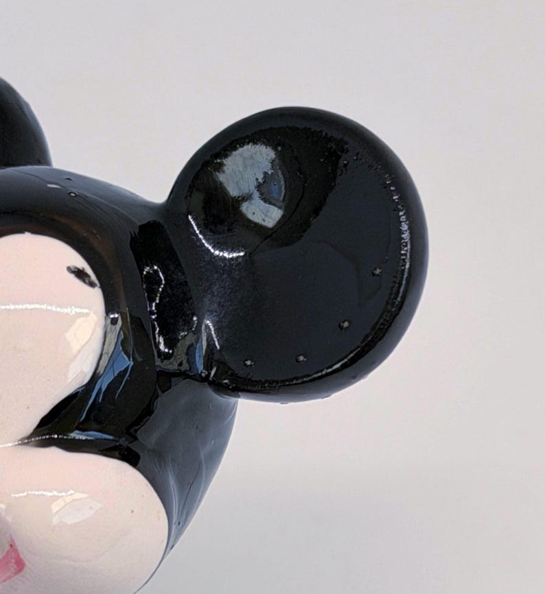 Puede incluir: Primer plano de una figura de Mickey Mouse en blanco y negro. La figura tiene un acabado brillante, con una gran oreja negra y una parte de la cara visible. La cara es blanca con acentos rosas.