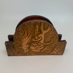May include: A copper napkin holder with a hammered finish, featuring a raised design of a person playing a guitar in front of a cactus. The holder has a wooden base.