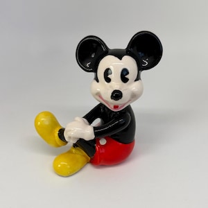 Op de afbeelding: Een keramieken beeldje van Mickey Mouse in een zittende positie. Het beeldje heeft zwarte oren en een zwart lichaam, witte handschoenen, een rode broek en gele schoenen. Het beeldje lacht en heeft een wit gezicht.