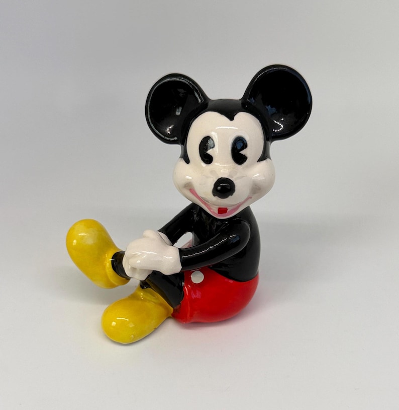 Op de afbeelding: Een keramieken beeldje van Mickey Mouse in een zittende positie. Het beeldje heeft zwarte oren en een zwart lichaam, witte handschoenen, een rode broek en gele schoenen. Het beeldje lacht en heeft een wit gezicht.