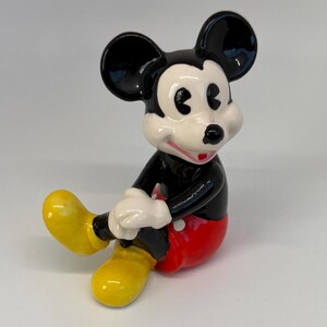 Op de afbeelding: Een keramieken beeldje van Mickey Mouse, met zwarte oren, een zwart lichaam, een rode broek en gele schoenen. Het beeldje heeft witte handschoenen en een lachend gezicht met een rode mond. Het klassieke Disney-personage zit.