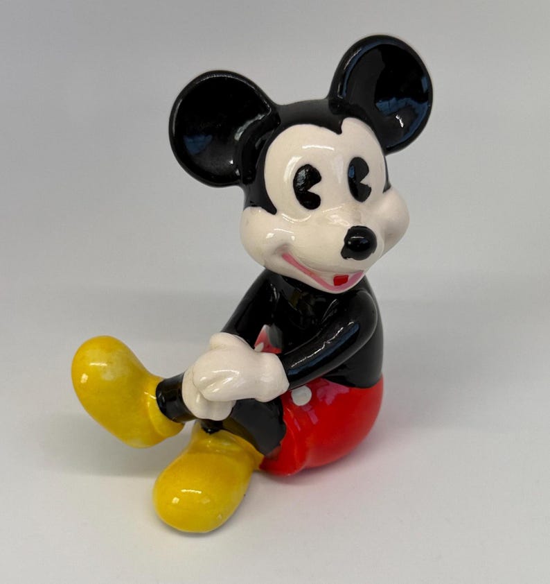 Op de afbeelding: Een keramieken beeldje van Mickey Mouse, met zwarte oren, een zwart lichaam, een rode broek en gele schoenen. Het beeldje heeft witte handschoenen en een lachend gezicht met een rode mond. Het klassieke Disney-personage zit.
