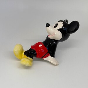 Puede incluir: Una figura de cer&aacute;mica de Mickey Mouse en una pose juguetona. La figura presenta un cuerpo negro, una cara blanca, pantalones cortos rojos y zapatos amarillos. El personaje cl&aacute;sico de Disney se representa con una expresi&oacute;n alegre.
