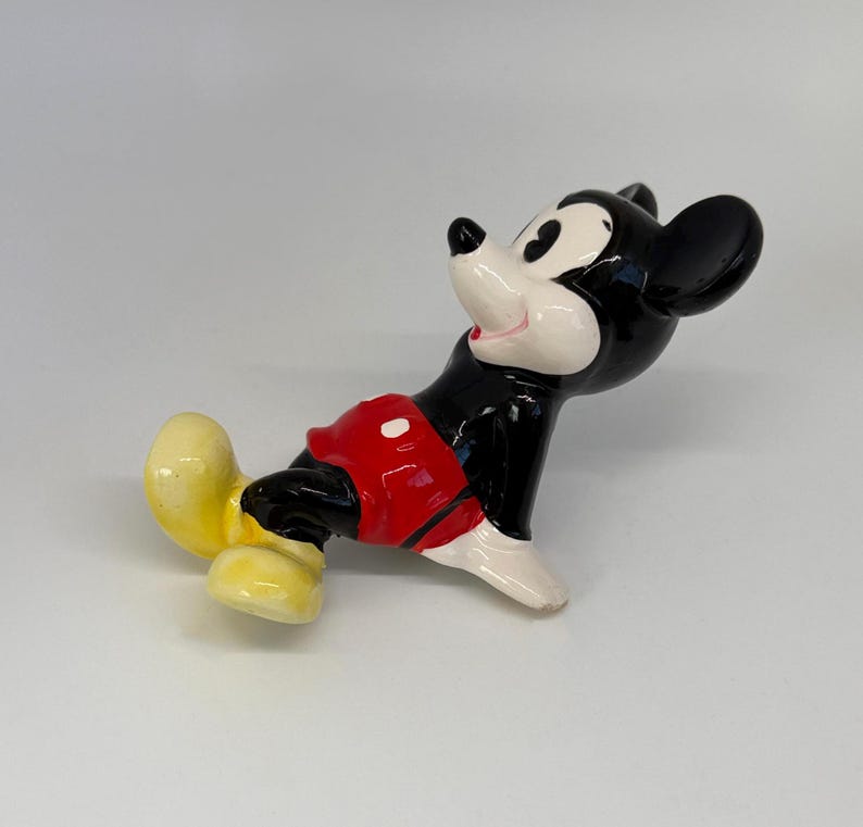 Puede incluir: Una figura de cer&aacute;mica de Mickey Mouse en una pose juguetona. La figura presenta un cuerpo negro, una cara blanca, pantalones cortos rojos y zapatos amarillos. El personaje cl&aacute;sico de Disney se representa con una expresi&oacute;n alegre.