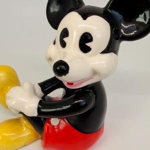 Op de afbeelding: Een keramieken Mickey Mouse beeldje, met een zwart lichaam, grote zwarte oren en een wit gezicht. Het beeldje draagt een rode broek en gele schoenen. Een verzamelobject.