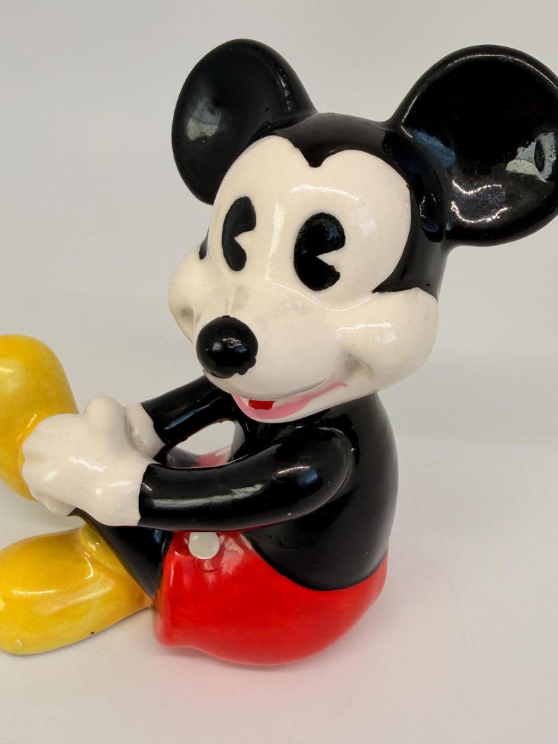 Op de afbeelding: Een keramieken Mickey Mouse beeldje, met een zwart lichaam, grote zwarte oren en een wit gezicht. Het beeldje draagt een rode broek en gele schoenen. Een verzamelobject.