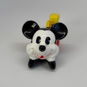 Puede incluir: Una figura de Mickey Mouse de cer&aacute;mica. La figura tiene una cara blanca, orejas negras y zapatos negros. Tiene un cuerpo rojo y un sombrero amarillo. La figura es un objeto de colecci&oacute;n.